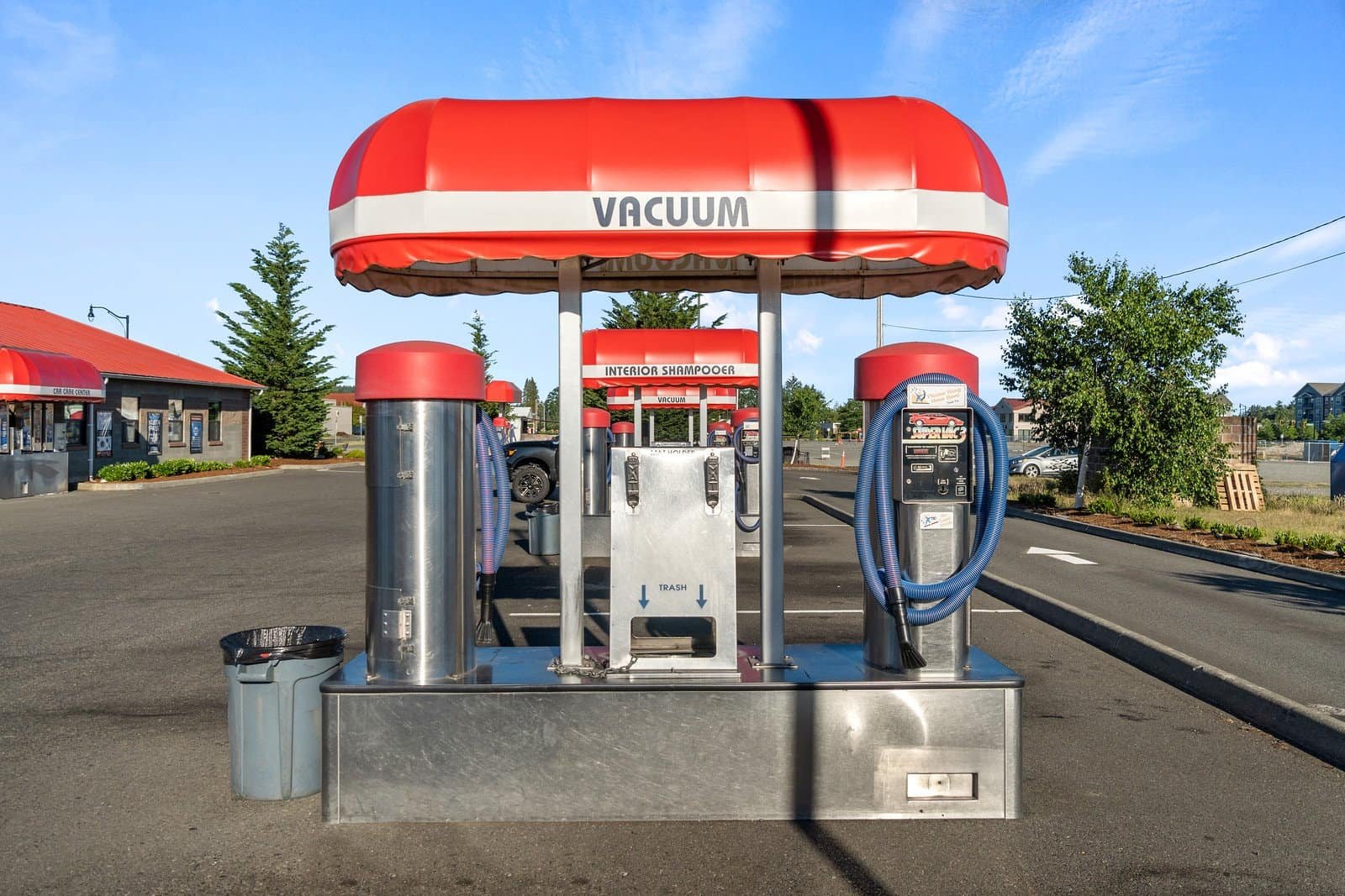A red and white vacuum station for cars stands in a parking lot, equipped with hoses and a coin-operated panel, under a clear blue sky. Trees, buildings, and a trash can are visible in the background.