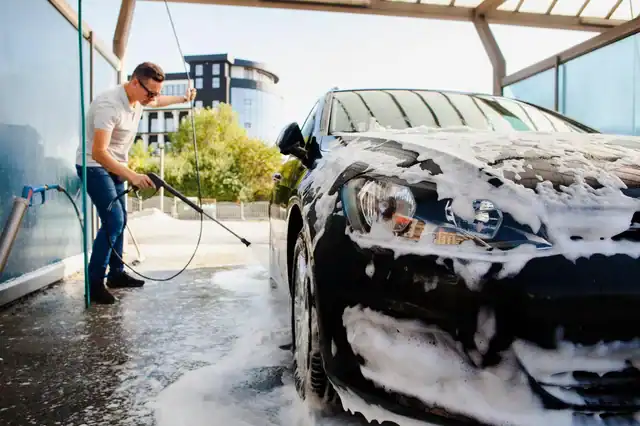 A man wearing sunglasses uses a high-pressure hose to wash a black car covered in soap suds at an outdoor car wash on a sunny day.