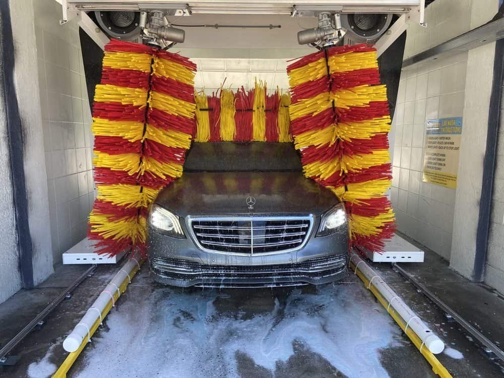 A black Mercedes-Benz inside an automatic car wash, with large spinning red and yellow brushes and soap suds covering the vehicle.